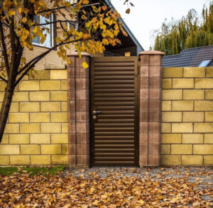 A modern, dark brown louvered metal gate is installed between two decorative stone-block pillars within a tan brick wall, with golden autumn leaves from a nearby tree scattered on the paved walkway.