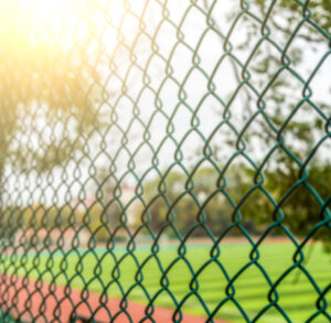 An intentionally blurred, close-up photo of a green chain-link fence with a bright sun flare in the upper left corner. Through the fence, a green athletic field and a red running track are visible in the out-of-focus background.