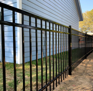 An angled view of a modern, flat-top black ornamental metal fence installed in a backyard. The fence, featuring vertical pickets, runs alongside a house with light blue siding, separating a green lawn from a dirt path.