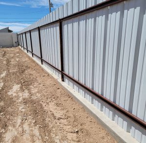 An angled view of a long privacy fence constructed with vertical, white corrugated metal panels and a dark brown metal frame. The fence is set on a concrete curb foundation, bordering a patch of dirt on a sunny day.